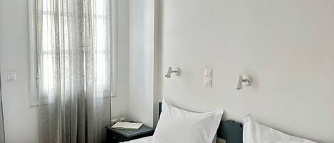 A neatly made bed with white linens and towels, a bedside table with a book, and a window with curtains.