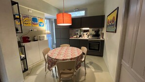 A compact kitchen with a dining area, featuring a round table with chairs, a hanging pendant light, and a window above the sink. - The houses of the Piazzetta - Apartment Levanzo (Trapani)