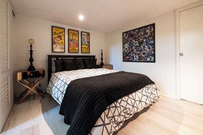 Ein Schlafzimmer mit einem Bett, einem Nachttisch, einer Lampe, einem Poster und einem Gemälde.