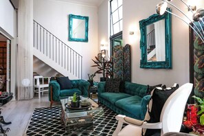 Ein Wohnzimmer mit türkisfarbenen Sofas, einem weißen Sessel, einer Treppe und spiegeln mit türkisnem Rahmen.