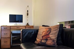 Ein Zimmer mit Schreibtisch, Computerbildschirm, Lampe und einer Couch mit Kissen.
