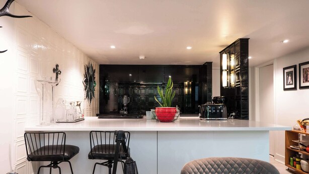 A modern kitchen with a white countertop, black bar stools, a red plant, and a black coffee machine.