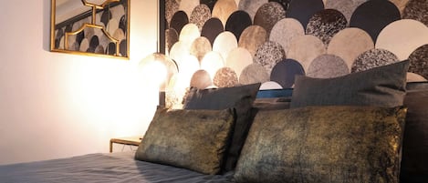 A bed with a headboard featuring a geometric pattern, a bedside table with a lamp, and a framed artwork on the wall.