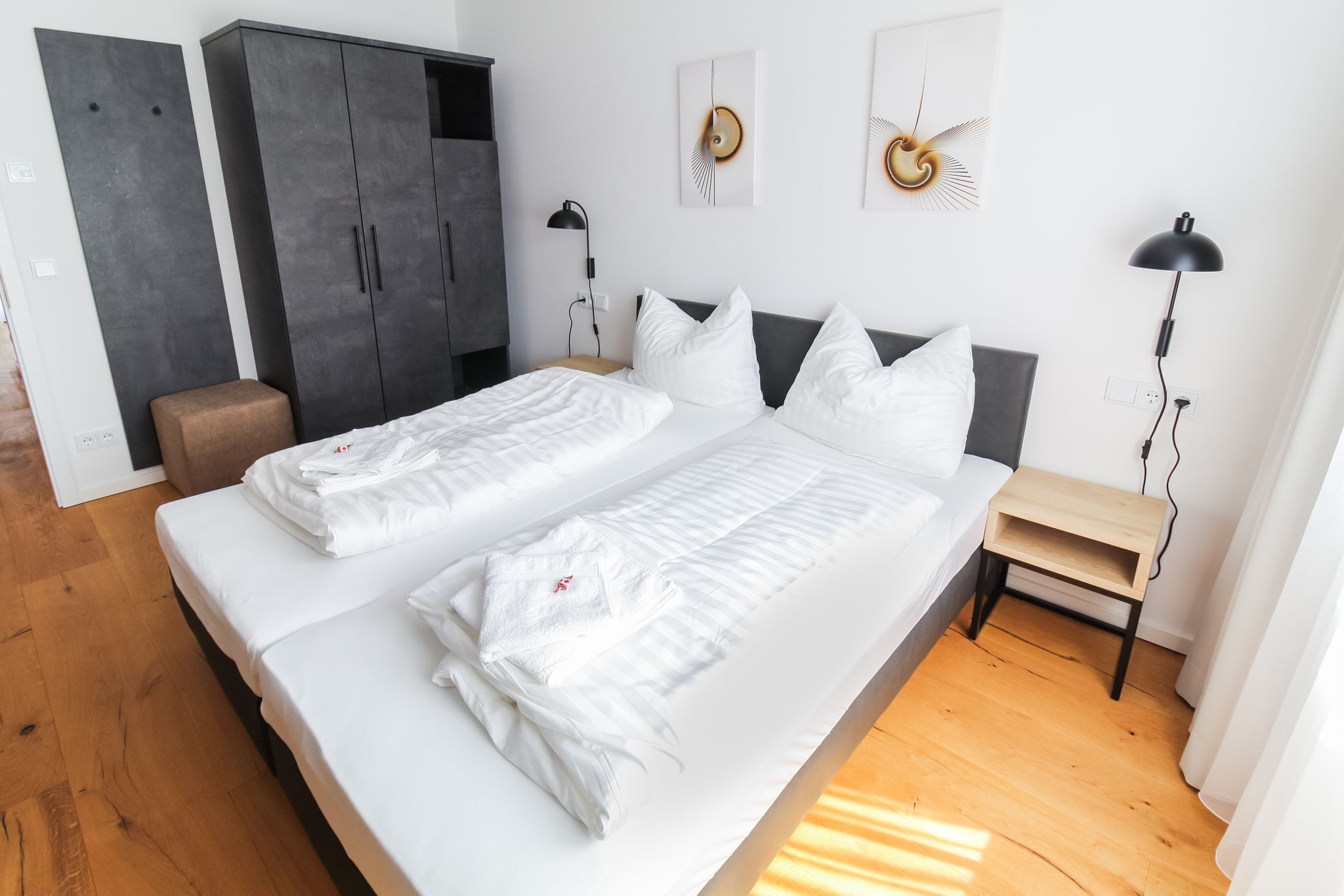 A double bed with white bedding, a wooden nightstand, a desk lamp, and a wardrobe.