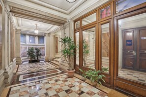 Un pasillo con piso de mármol a cuadros, plantas grandes en macetas y una puerta de madera con un letrero.