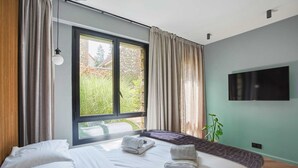 A modern bedroom with a large window, a bed with white linens, a flat-screen TV mounted on the wall, and a potted plant. - Superb Suite in Neuilly Porte Maillot (Neuilly-sur-Seine)