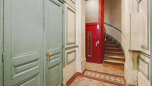 A narrow hallway with a red door and a staircase leading to an upper floor. The walls are painted in a light color, and the floor has a patterned tile design. - Modern Studio with AC in the heart of Paris (Paris)