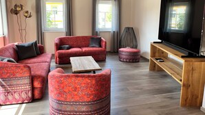 Un salon avec un canapé rouge, un pouf assorti, une table basse et un téléviseur posé sur un meuble en bois. Il y a deux fenêtres avec des rideaux, une lampe sur pied et un luminaire fixé au mur.