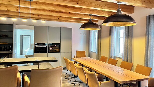 Une cuisine moderne dotée d’appareils électroménagers intégrés, un coin repas avec une longue table en bois et des chaises assorties, et un plafond orné de poutres apparentes et agrémenté de luminaires suspendus.