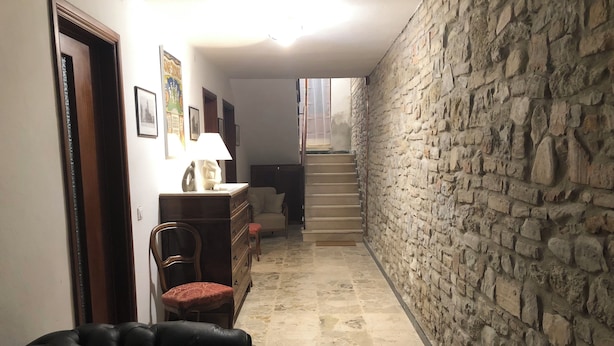 A narrow hallway with a stone wall on one side and a wooden door on the other. There is a chair and a small table with a lamp in the center. A staircase leads to an upper level. - Residence "The Castle" 1  (Castell'Arquato)