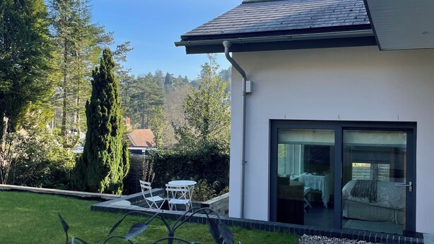 Una casa moderna con un ventanal de vidrio que da a un dormitorio, un pequeño patio con mesa y sillas, y un césped bien cuidado.