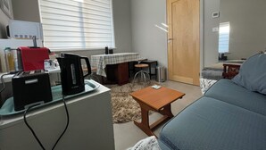 Una sala de estar con un sofá, una mesa de centro, una mesa con mantel a cuadros, una ventana con persianas, una puerta y una zona de kitchenette con una cafetera Bosch y un electrodoméstico rojo.