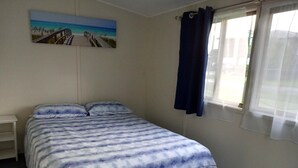 A bedroom with a bed, a nightstand, a painting of a beach, and a window with curtains. - Perfect Fishing Holiday (Burrum Heads)