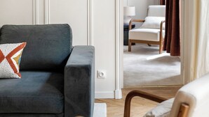 A modern living room with a dark grey sofa, a wooden chair, and a glass coffee table. A mirror reflects a room with a white chair and a lamp. - Trevise (Paris)