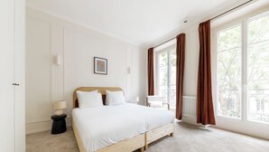 A bedroom with a large bed, a nightstand, a chair, and a window with curtains. - Trevise (Paris)