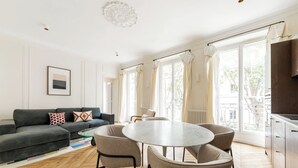 A modern living room with a grey sofa, a round table with chairs, and large windows with curtains. - Trevise (Paris)
