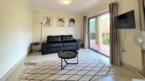 A living room with a black sofa, a small side table, a coffee table, a TV mounted on the wall, and a sliding glass door leading to an outdoor area. - The Sandcastle at Burrum Heads (Burrum Heads)