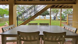 An outdoor patio with a wooden table and chairs, a metal staircase, and a view of a grassy area and trees. - The Sandcastle at Burrum Heads (Burrum Heads)