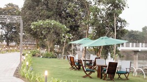 An outdoor seating area with tables and chairs, surrounded by greenery and a body of water. - Praveg Rezort Kachigam (Daman)