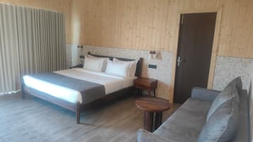 A hotel room with a wooden paneled wall, a bed with white bedding, a grey sofa, a small wooden table, and a door.