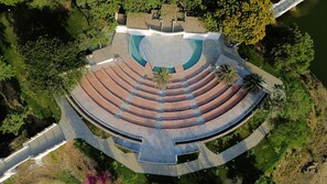 Eine Vogelperspektive eines kreisförmigen Amphitheaters mit gestuften Sitzreihen, umgeben von viel Grün und einem Wasserspiegel.