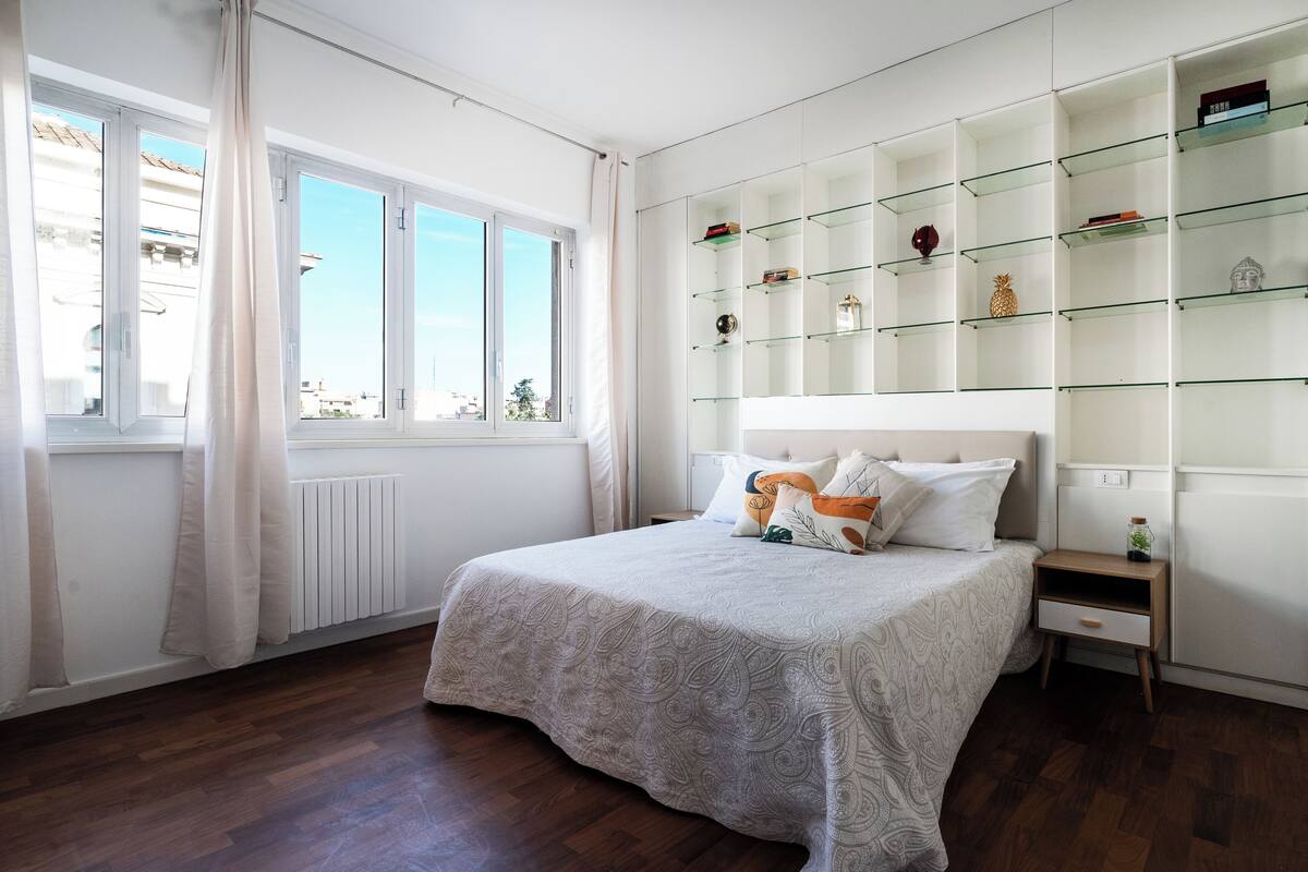 Un dormitorio con una cama grande, una mesita de noche, una estantería de vidrio empotrada y una ventana con cortinas.