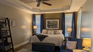 Une chambre à coucher comprenant un lit, une chaise, une table de chevet, une lampe, un ventilateur de plafond et un mur décoré d’œuvres d’art.