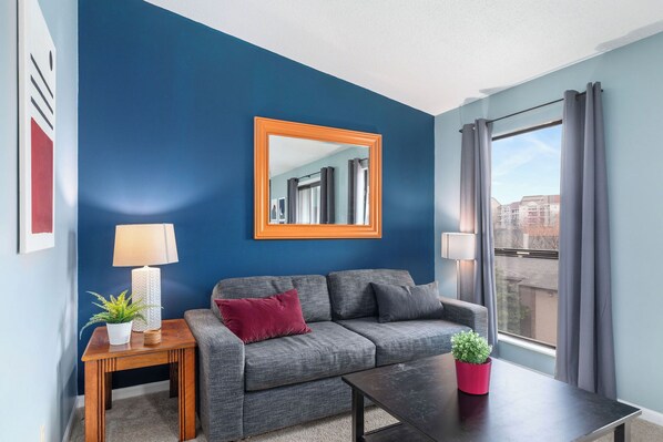 Una sala moderna con una pared azul de acento, un sofá gris, una mesa de centro de madera y un ventanal con cortinas.
