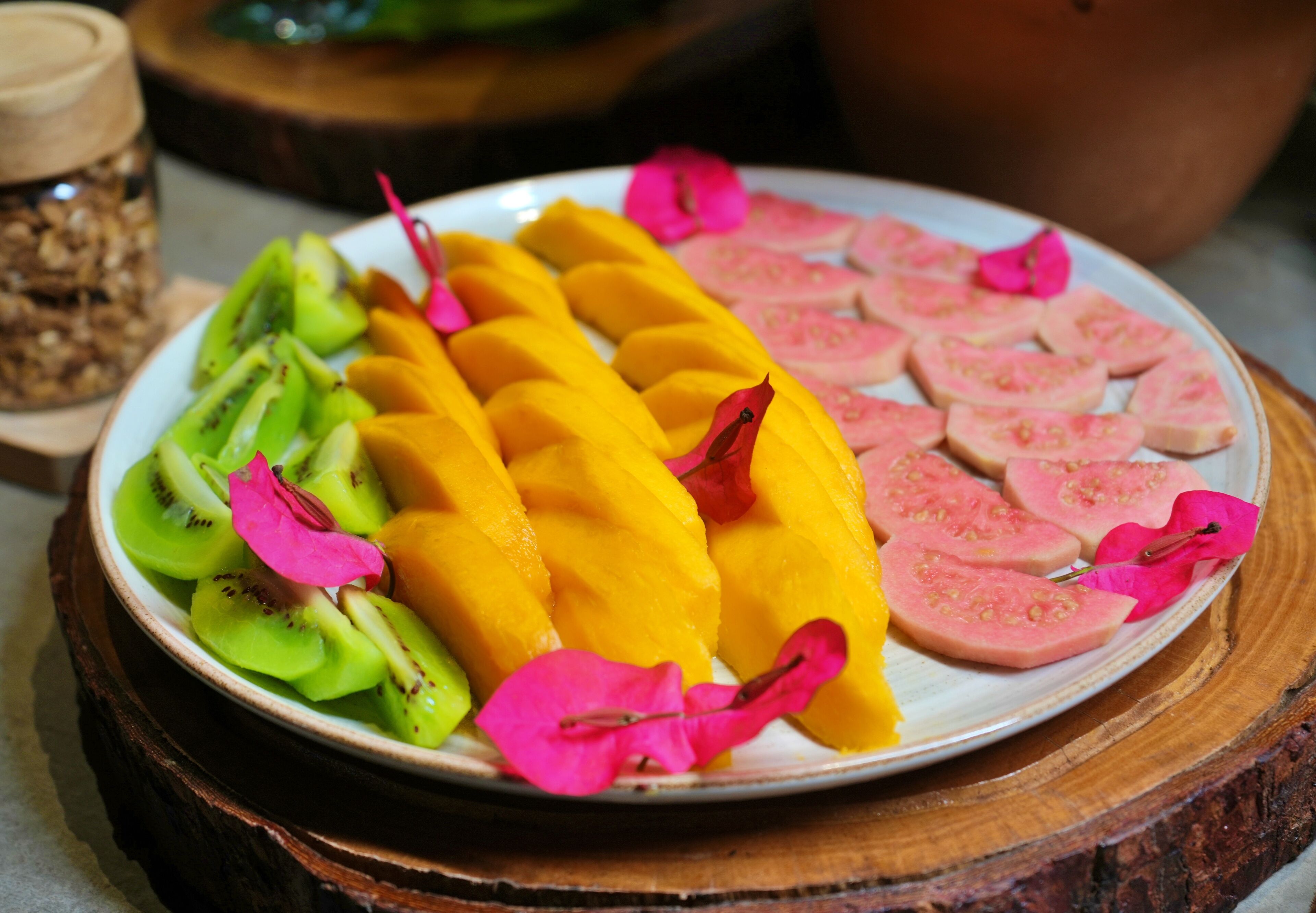 A plate with sliced mangoes, kiwi, and pink tomatoes, garnished with pink flowers.