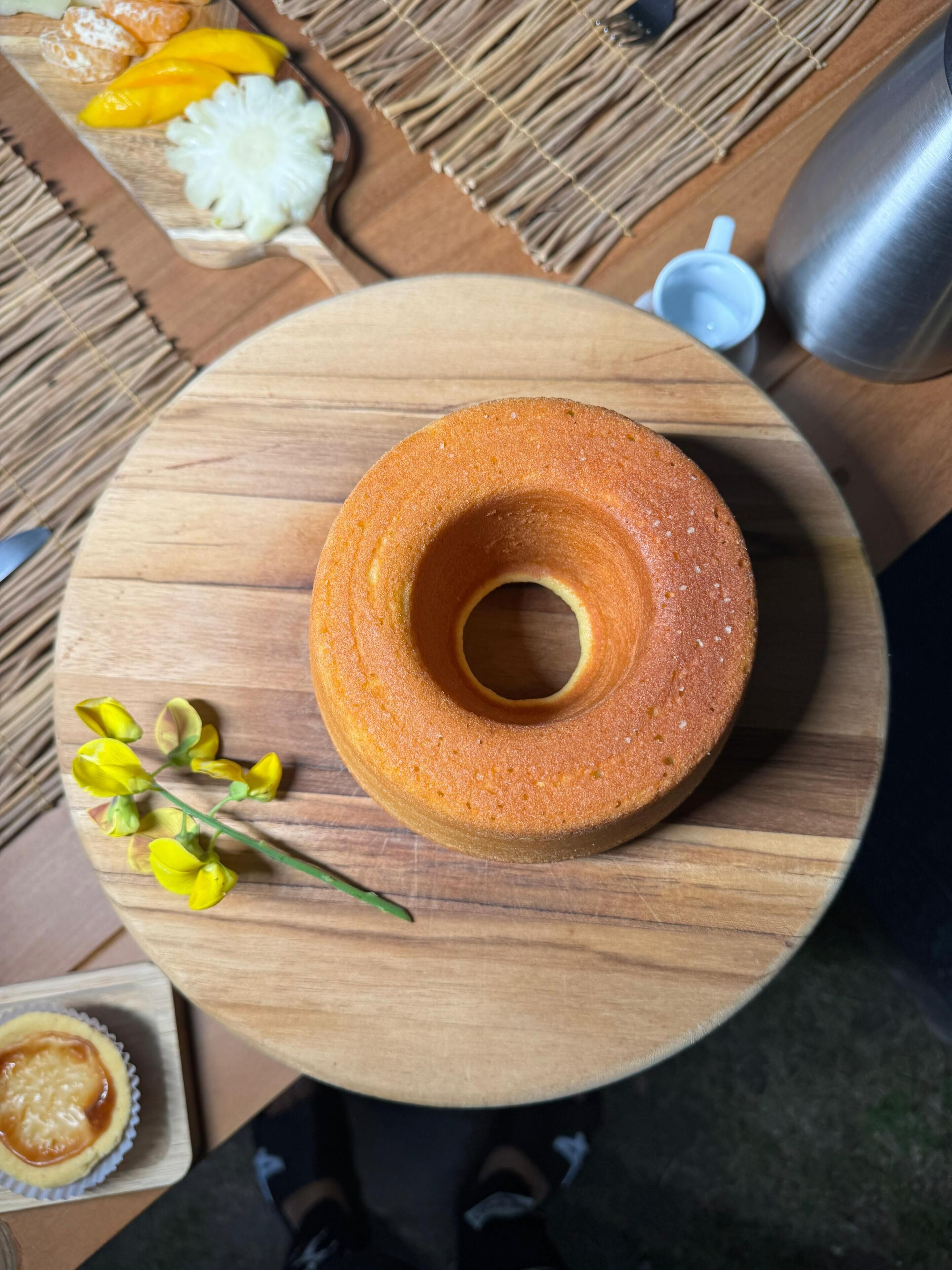 A round wooden table with a bundt cake, a small bowl of yellow flowers, and a metal pitcher.