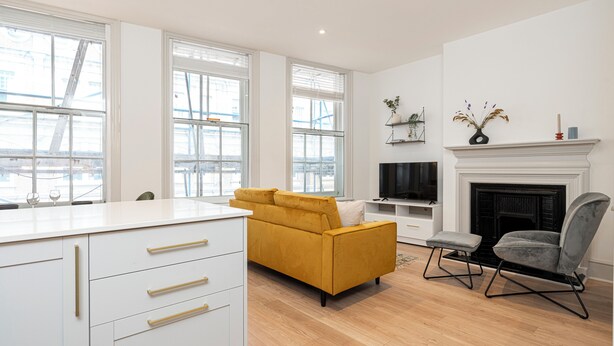A modern living room with a yellow sofa, a grey armchair, and a fireplace. - JOIVY Stylish 1 bed flats in Soho (London)