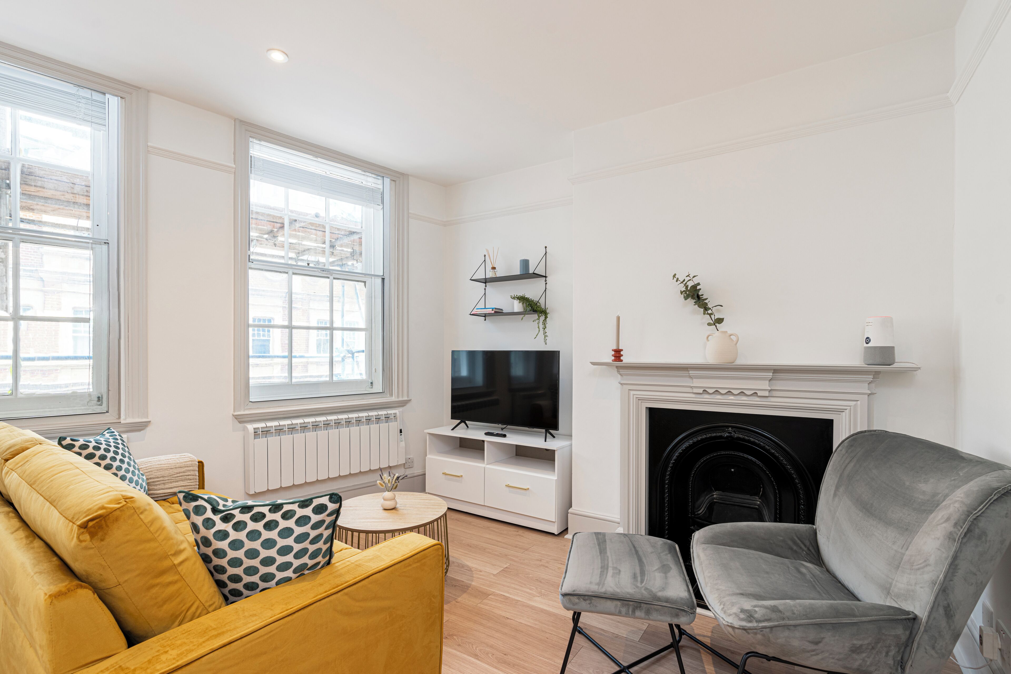 A living room with a yellow sofa, a grey armchair, a TV on a white stand, a fireplace, and large windows.