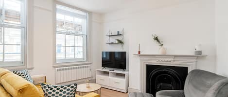 A living room with a yellow sofa, a grey armchair, a TV on a white stand, a fireplace, and large windows.