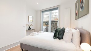 A modern bedroom with a large bed, a view of the outside, and a small table with a vase. - JOIVY Stylish 1 bed flats in Soho (London)