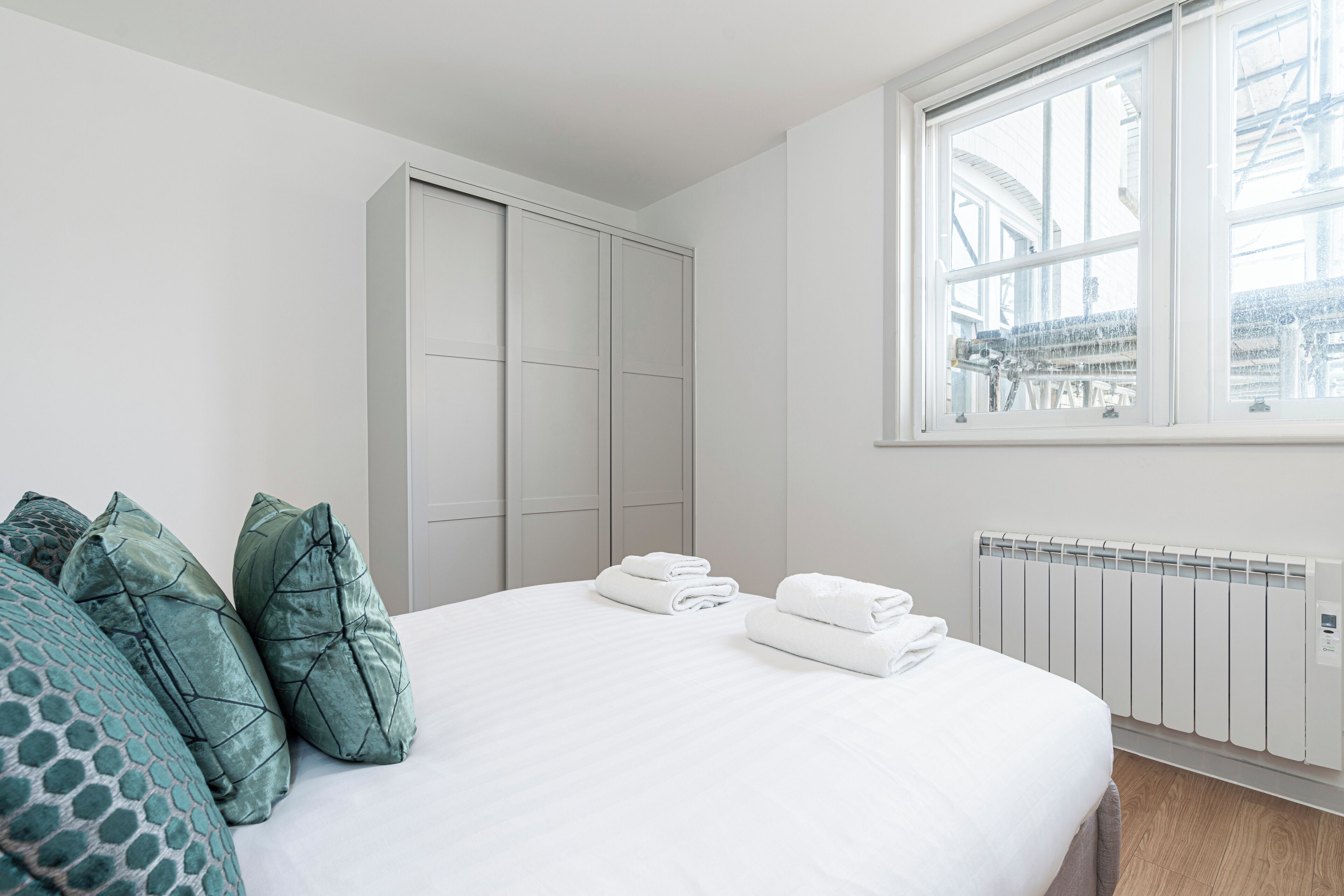A neatly made bed with pillows, a radiator, and a window with a view of buildings.