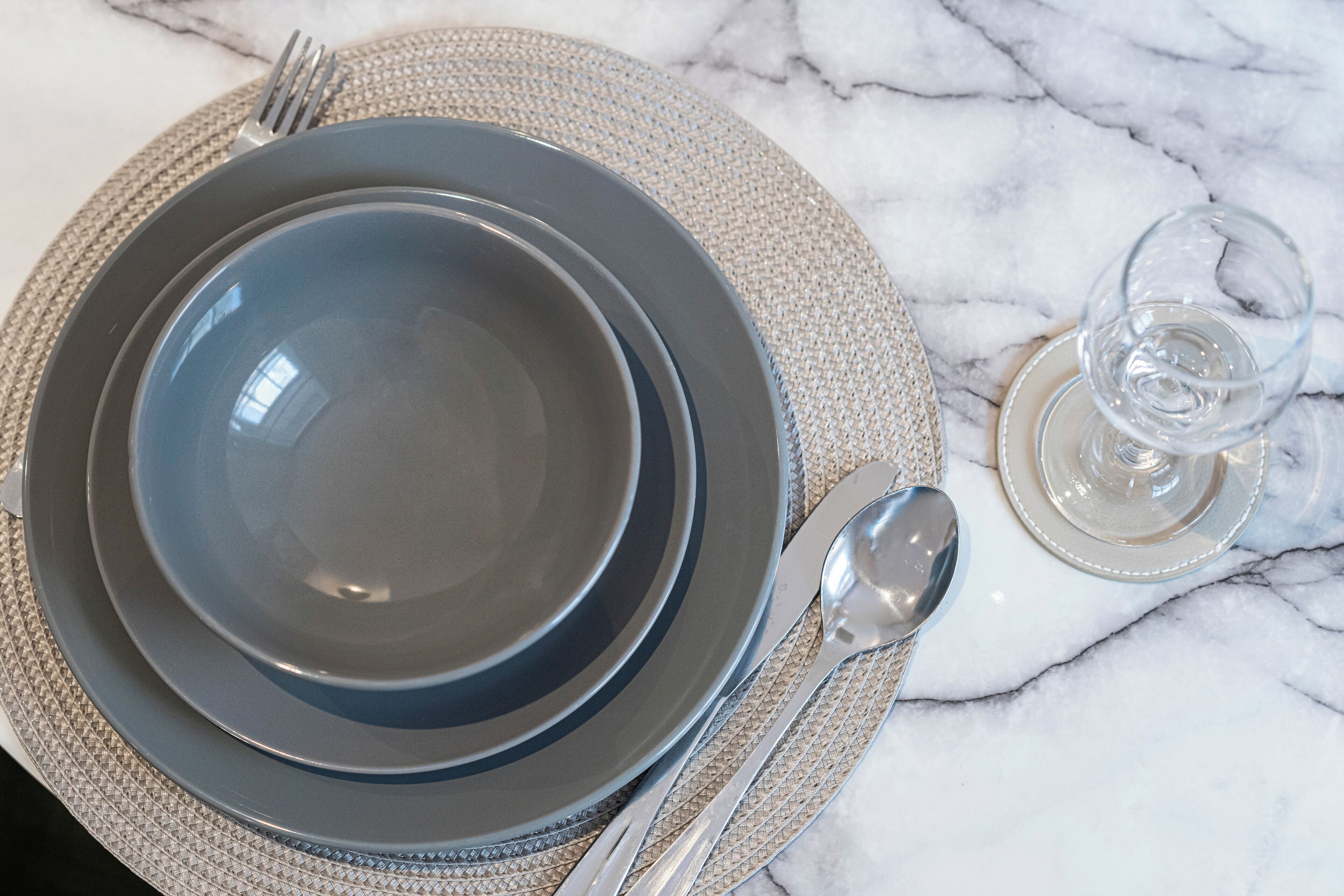 A table set with a dark plate, a fork, a spoon, and a glass of water on a marble surface.