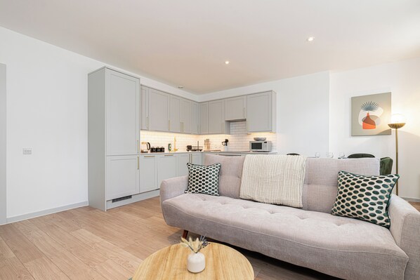 A modern living room with a grey sofa, a round coffee table, and a kitchen in the background. - JOIVY Stylish 1 bed flats in Soho (London)