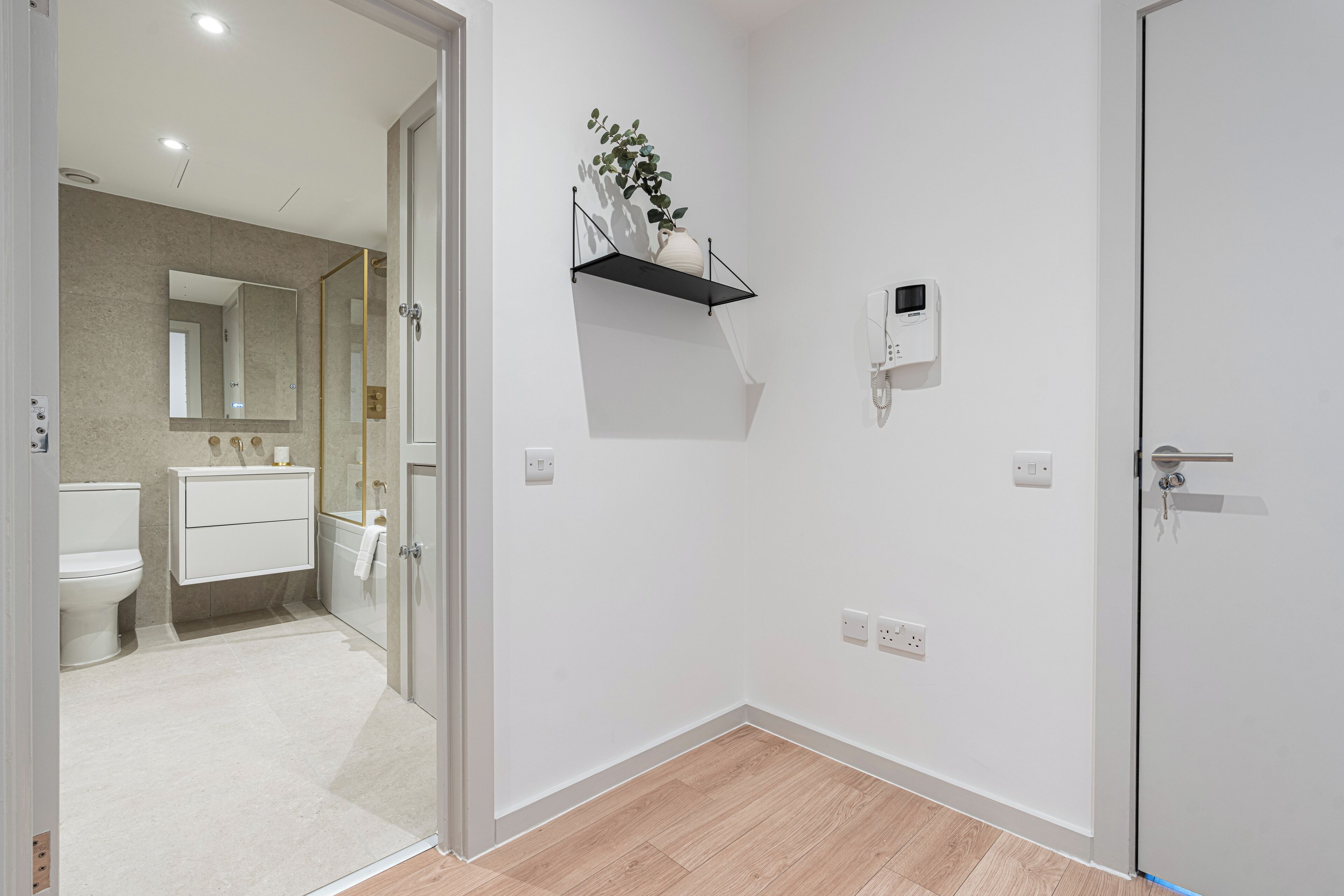 Uma casa de banho moderna com sanita, lavatório e espelho. Há uma prateleira com uma planta e um dispositivo fixado na parede.