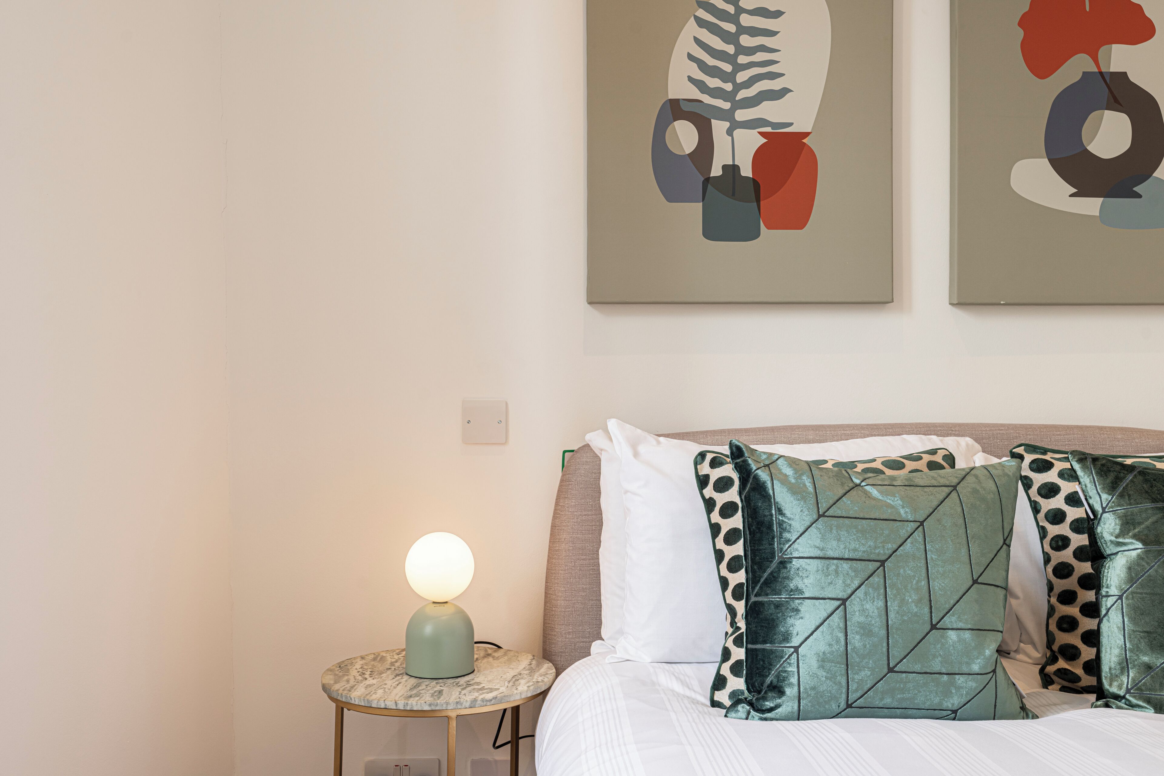 Een netjes opgemaakt bed met gestreepte kussens, een rond nachtkastje met een lamp, en twee abstracte schilderijen aan de muur.