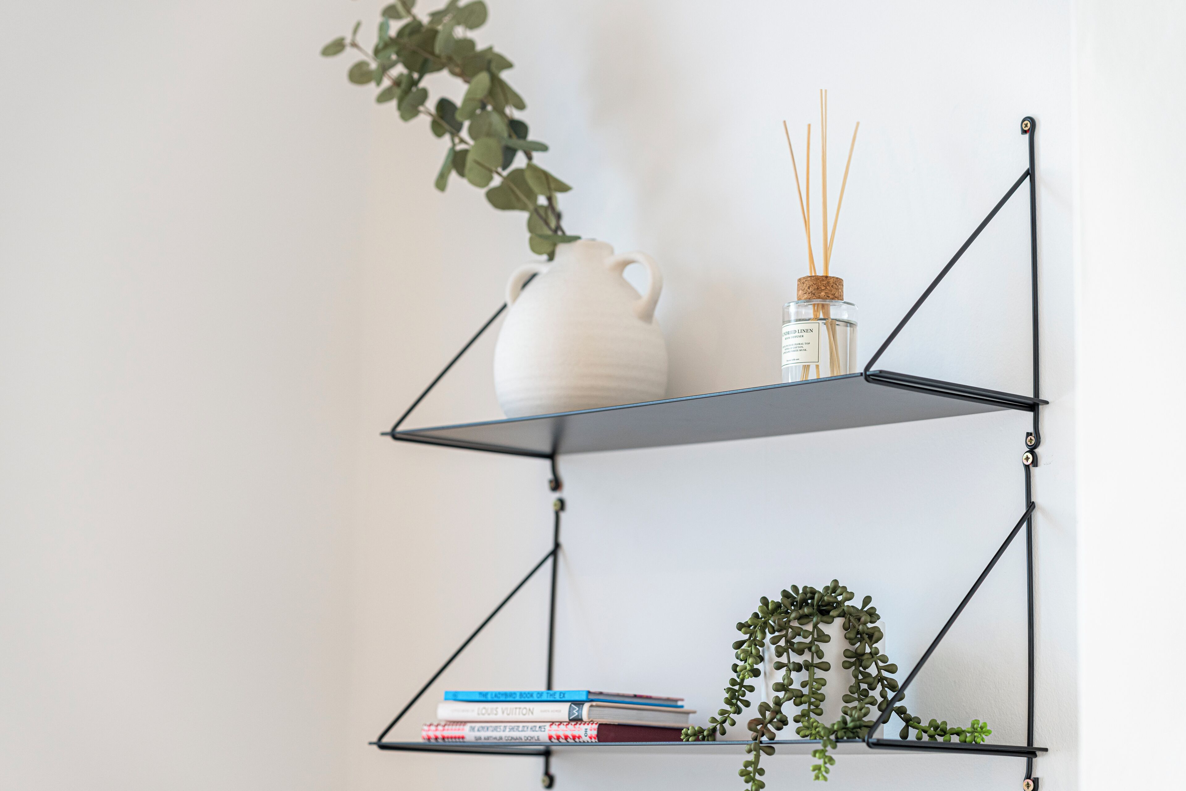 Una jarra de cerámica blanca sobre una estantería de metal negro con libros y una pequeña planta.