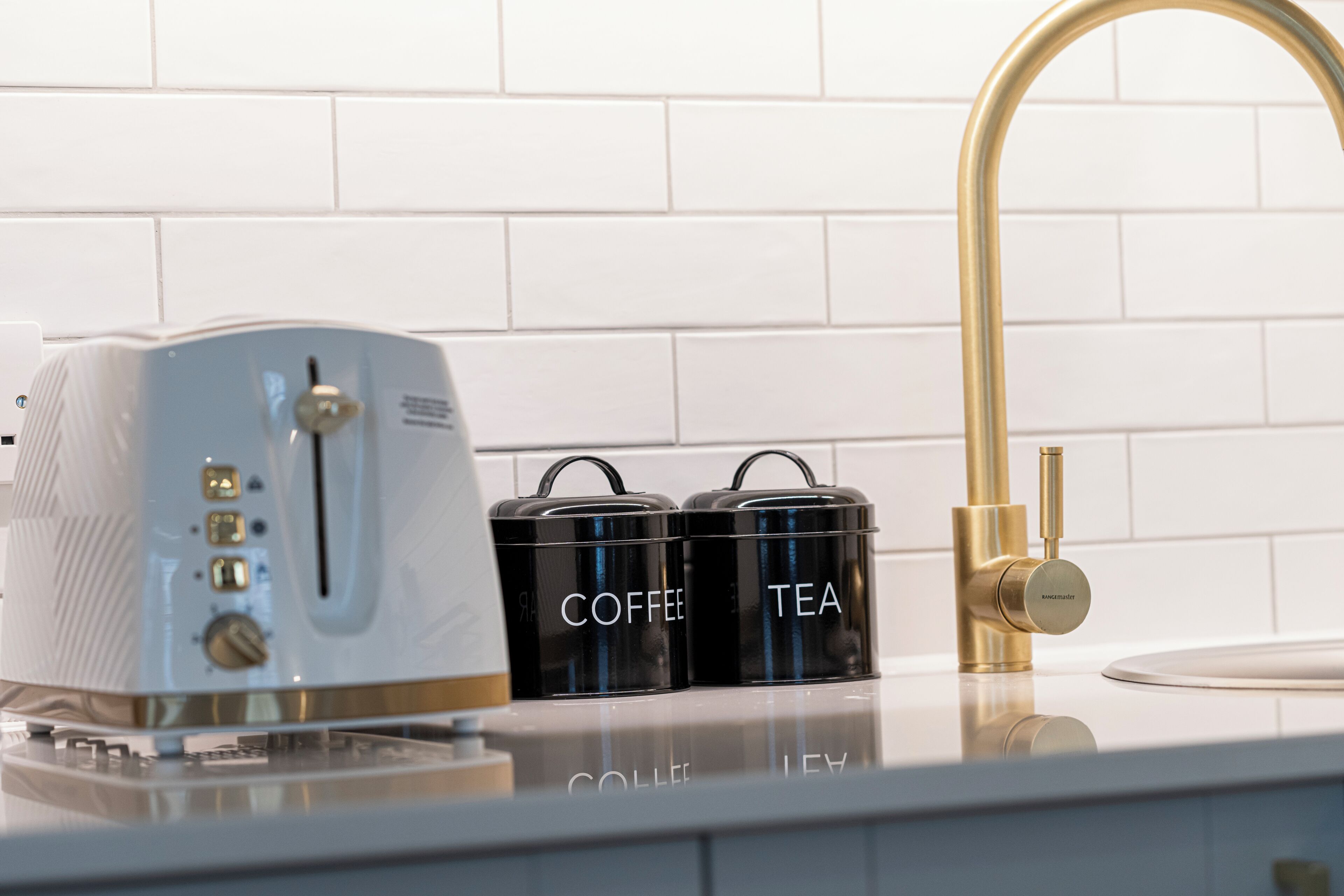 A kitchen with a white countertop, a white toaster, two black containers labeled "Coffee" and "Tea," and a gold faucet.