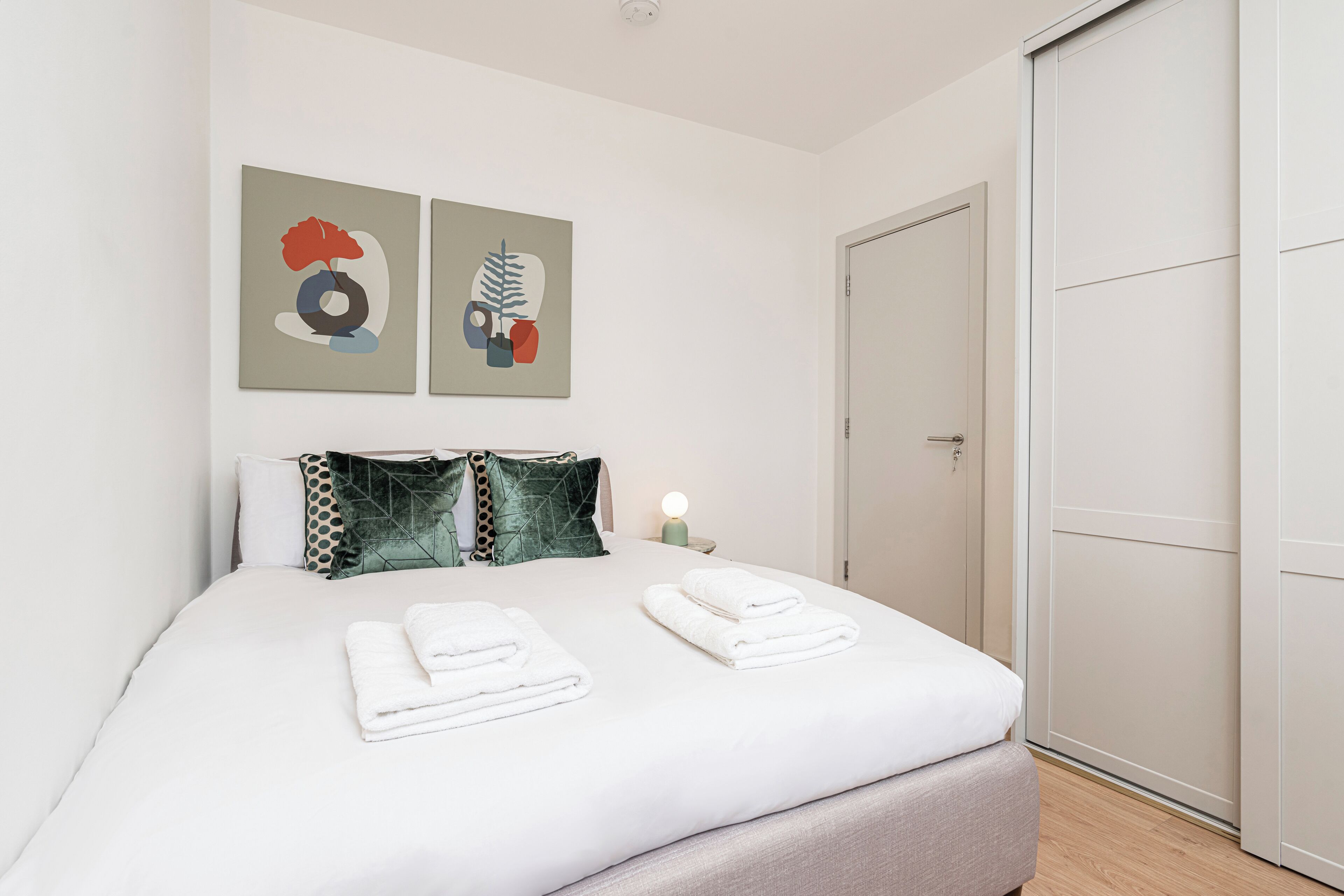 A neatly made bed with two folded towels, a bedside lamp, and two abstract wall art pieces.