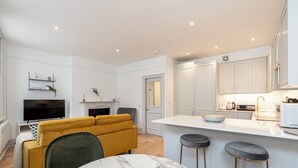 A modern kitchen with white cabinets, a marble countertop, and a yellow sofa. - JOIVY Stylish 1 bed flats in Soho (London)