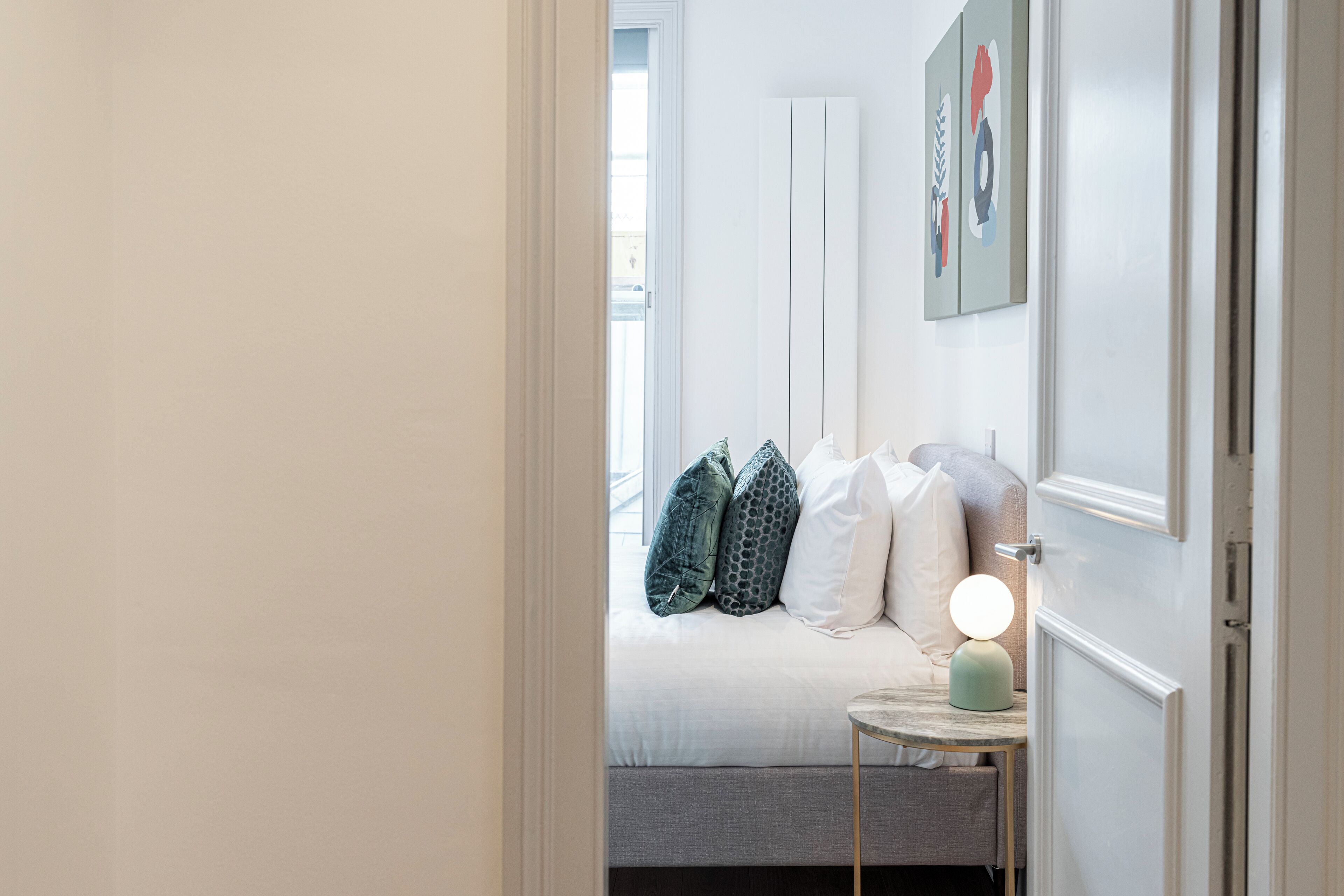 A bedroom with a bed, pillows, a bedside table with a lamp, and a door leading to another room.