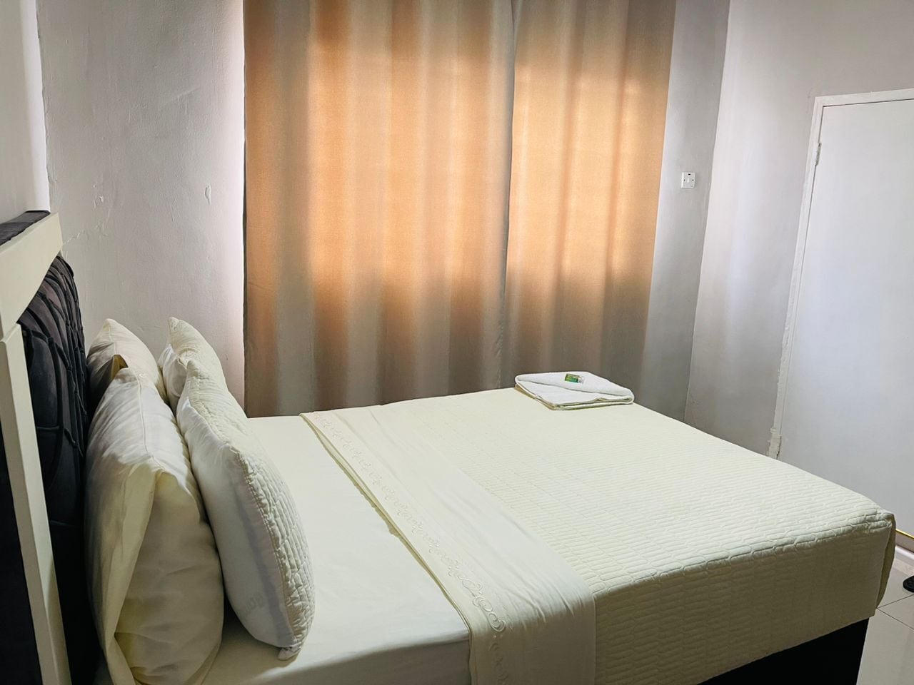 Una cama bien hecha con almohadas y una toalla doblada, una puerta blanca y una ventana con cortinas.