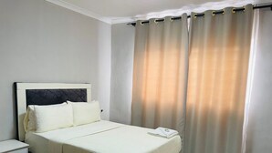 Una cama bien hecha con una colcha blanca y almohadas, una mesita de noche con una lámpara y una ventana con cortinas beige.
