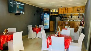 Un restaurante con mesas cubiertas de manteles rojos, sillas blancas, un refrigerador Vetra y un televisor mostrando un canal deportivo.