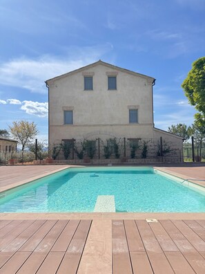A rectangular swimming pool with a wooden deck in the foreground and a large building with a flat roof and multiple windows in the background. - Relais La Colombaia (Appignano)
