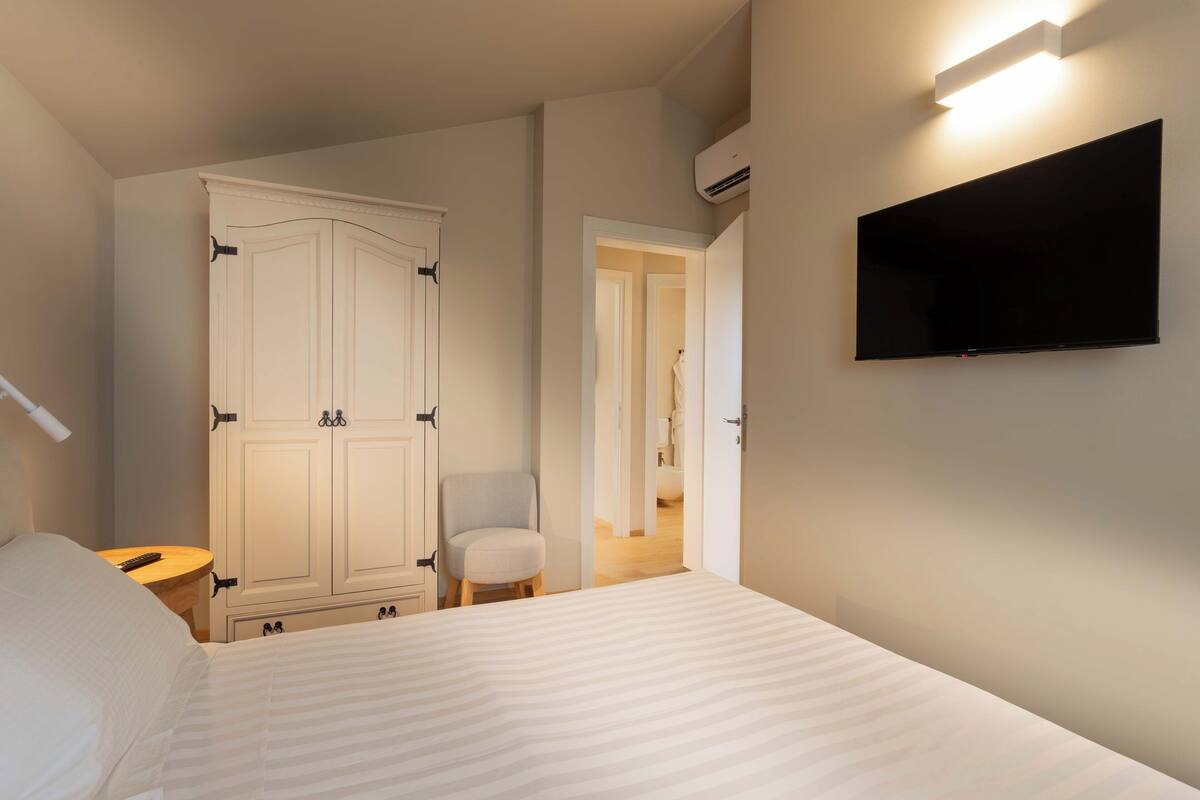 Un dormitorio con cama, armario, silla, televisor montado en la pared y una mesita de noche con lámpara.