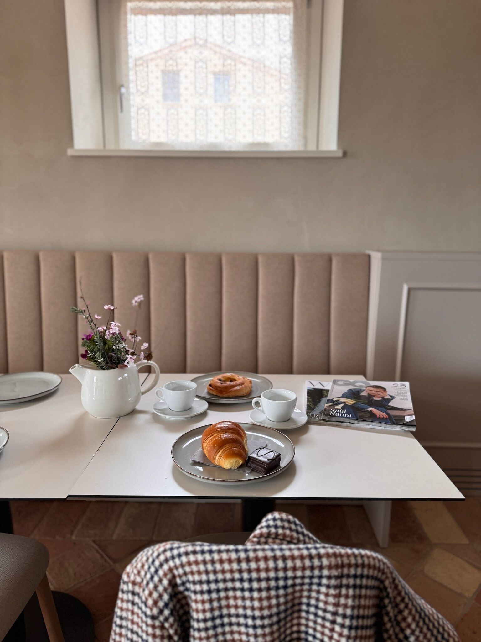 Une table à manger avec un pichet blanc, deux tasses, une assiette contenant un croissant et un morceau de chocolat, et un magazine.